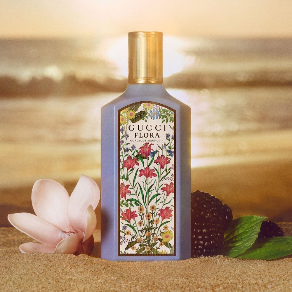 Flacone con etichetta floreale. Tappo dorato. Scritta: Gucci Flora Gorgeous Magnolia. Sfondo spiaggia.