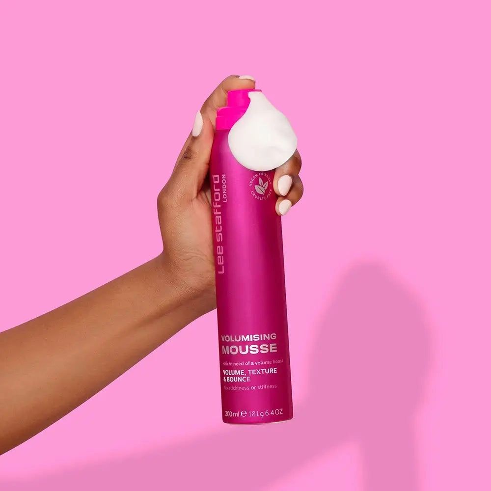 Mano che tiene una lattina rosa di Lee Stafford Volumising Mousse, con fuoriuscita di schiuma bianca.