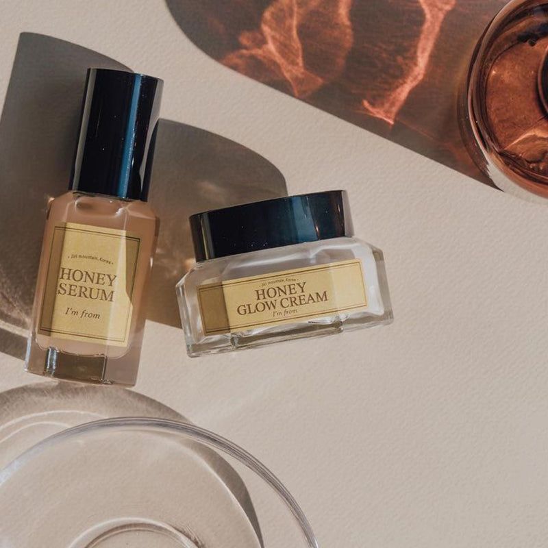 Flacone e vasetto di crema. Scritta: HONEY SERUM, HONEY GLOW CREAM, I'm from. Sfondo beige.