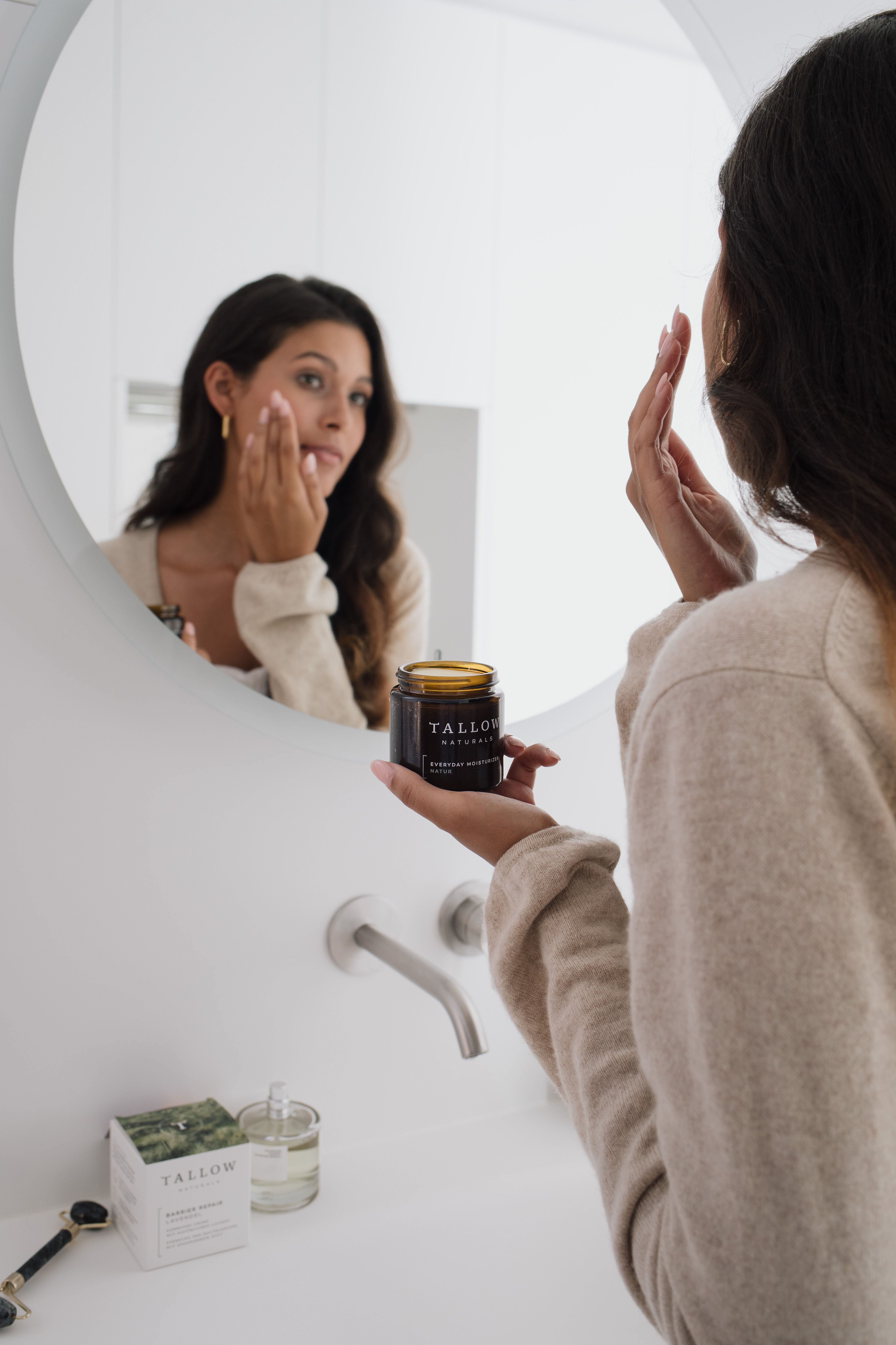 Donna si guarda allo specchio. Tiene un barattolo marrone con coperchio nero. Scritta: TALLOW NATURALS, BARRIER REPAIR LAVENDEL.