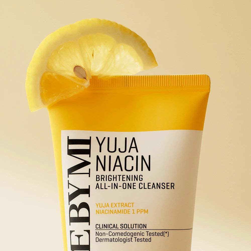 Tubo giallo con etichetta bianca. Scritta: Yuja Niacin Brightening All-in-One Cleanser. Fetta di limone come decorazione.