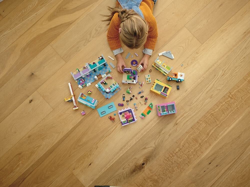 Un bambino gioca con i mattoncini LEGO su un pavimento di legno. Varie parti LEGO e minifigure sono sparse. Il bambino è visto dall'alto e si concentra sulla costruzione.