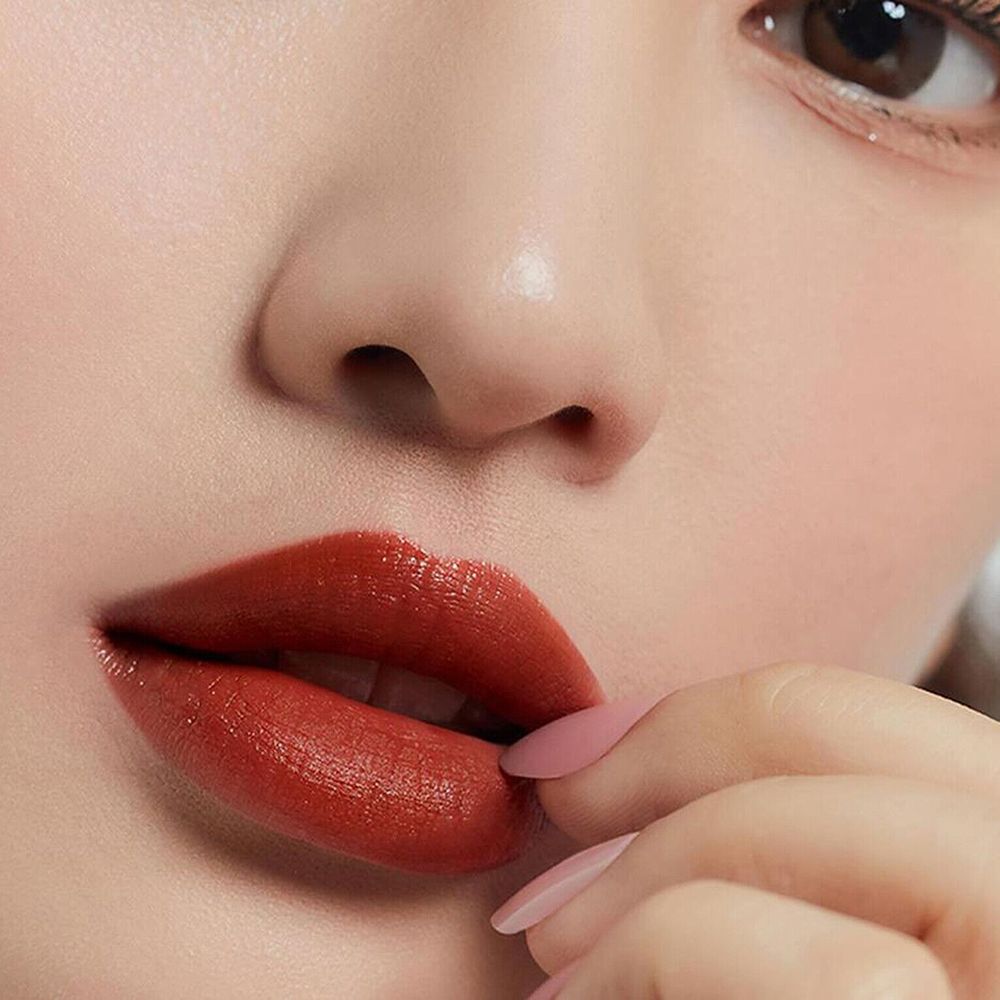 Primo piano di labbra con lucidalabbra marrone-rossastro. Una mano con unghie rosa tocca le labbra.