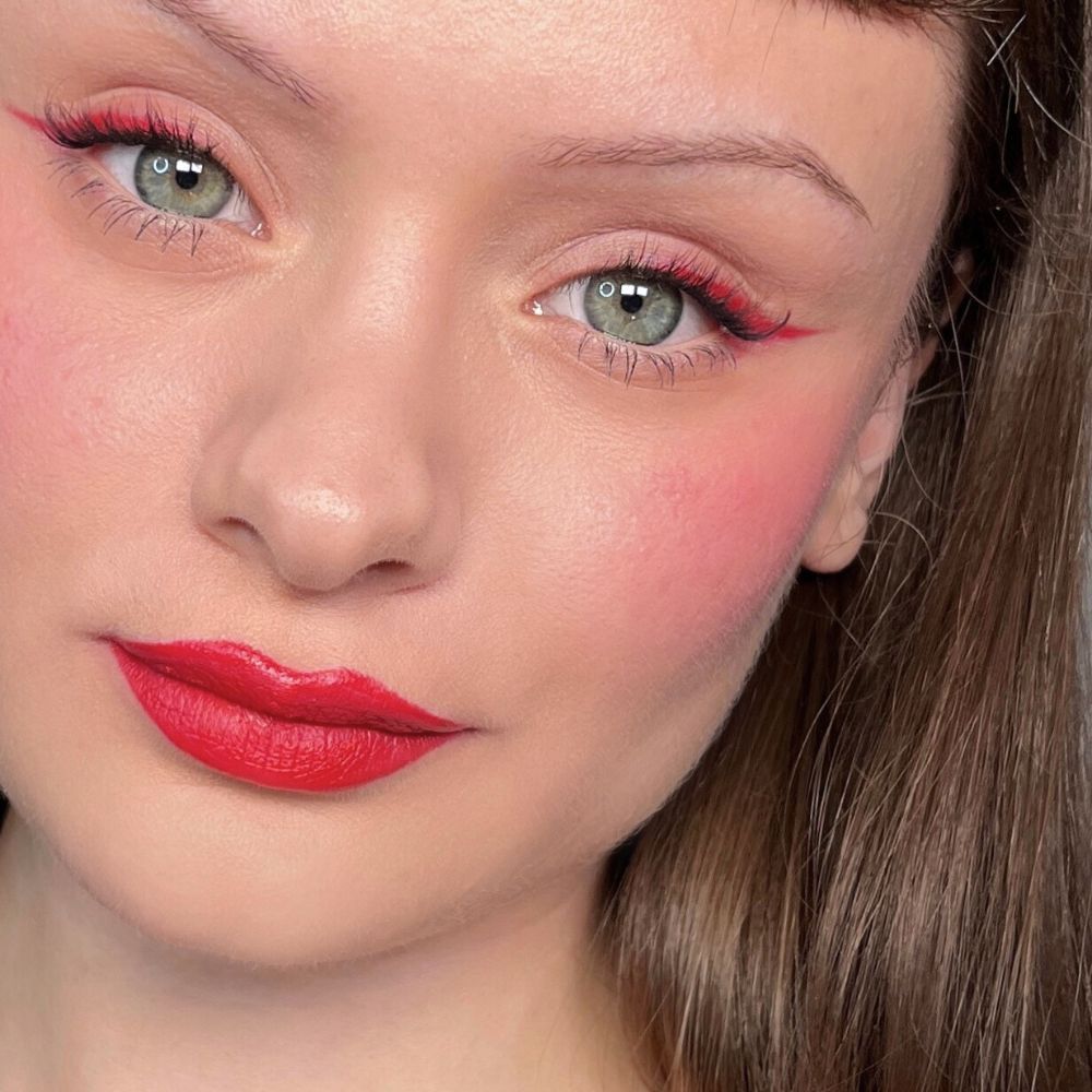 Primo piano di un viso con eyeliner, fard e rossetto rossi. Occhi verdi. Prodotto: ALL TIGERS.