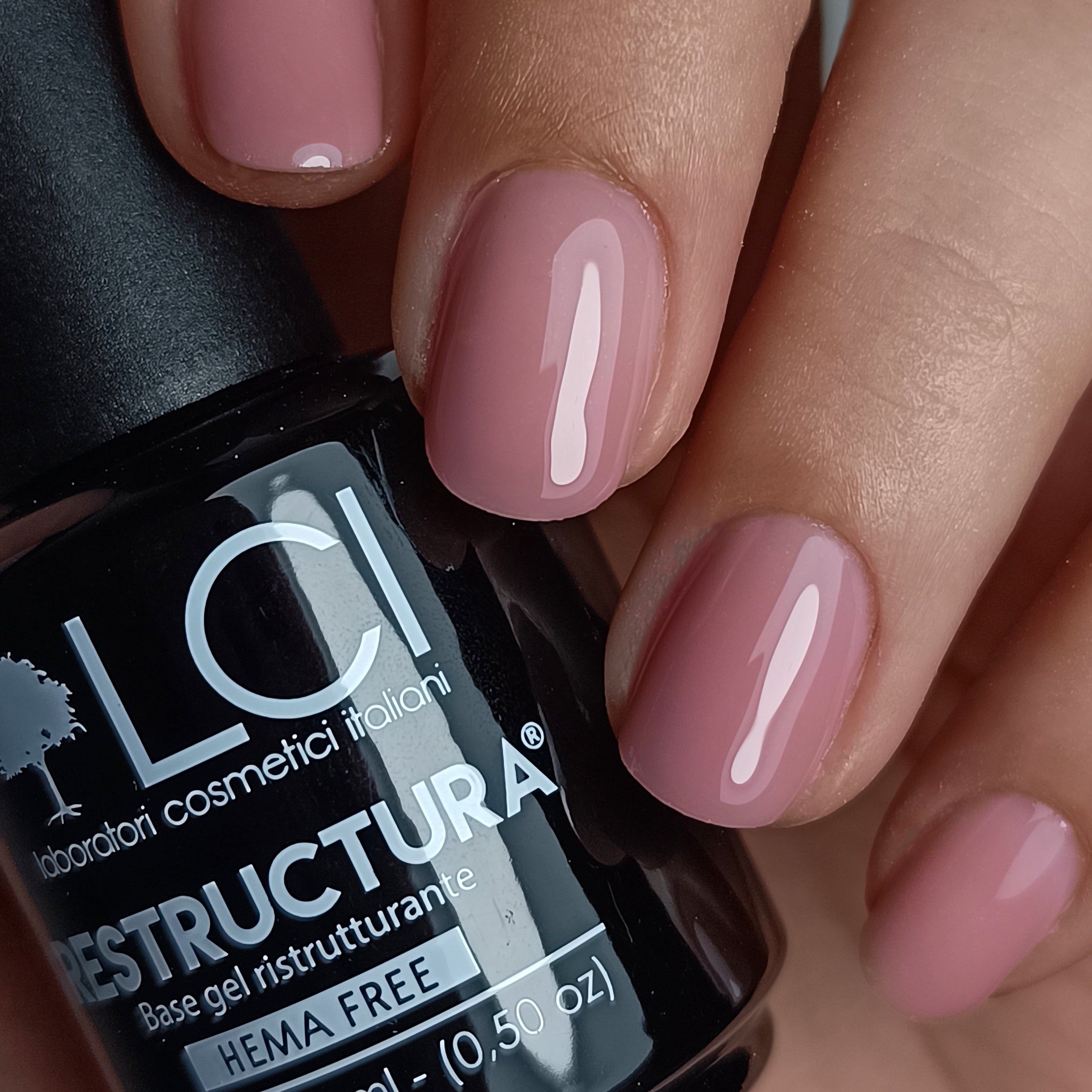 Mano con unghie smaltate di tonalità rosa. Flacone LCI Cosmetics Restructura Base&Go sullo sfondo.