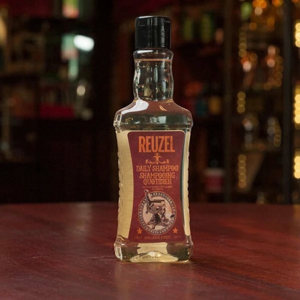 Piccolo flacone di shampoo. "Reuzel Daily Shampooing" sull'etichetta. Tappo nero. Posizionato su un tavolo.