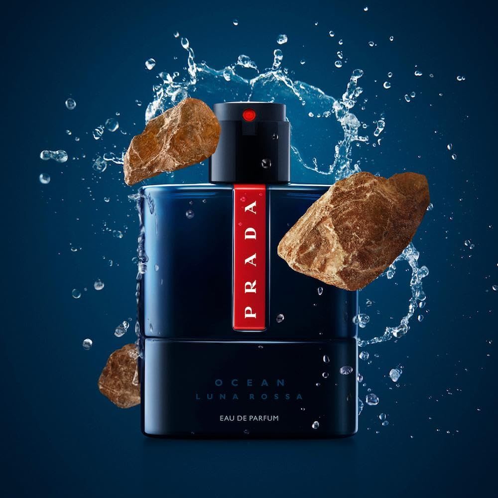 Flacone blu scuro con striscia rossa Prada. Erogatore nero. Pietre e spruzzi d'acqua.