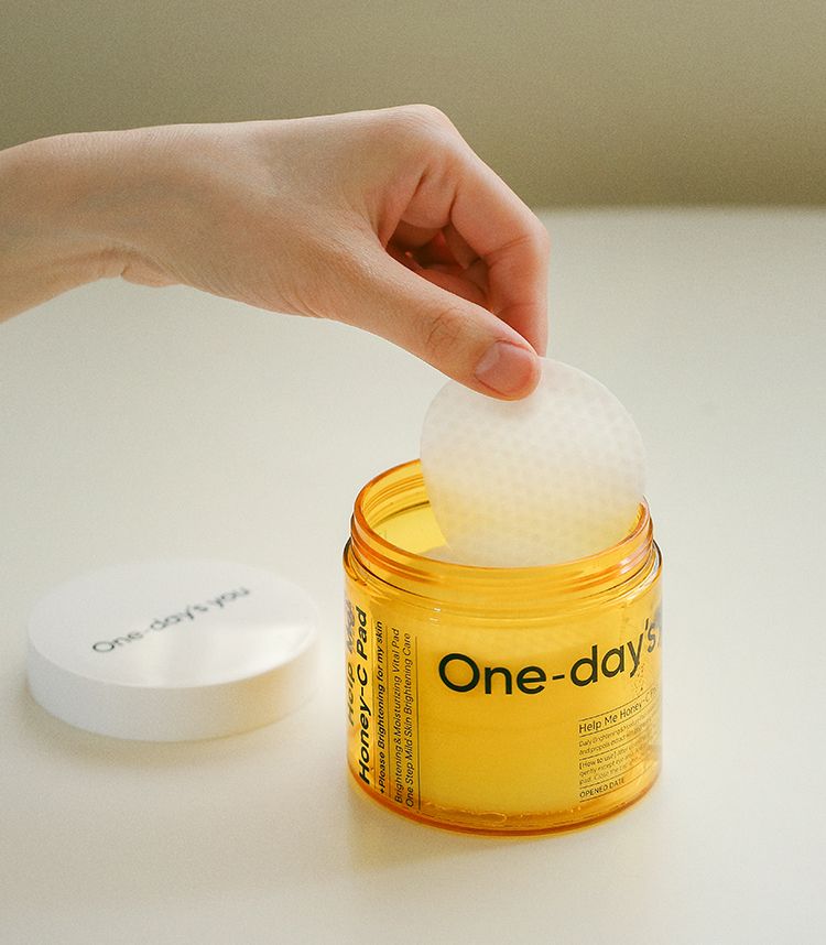 Un barattolo rotondo, aperto e giallo con la scritta "One-day's you Honey-C Pad". Una mano estrae un dischetto di cotone imbevuto.