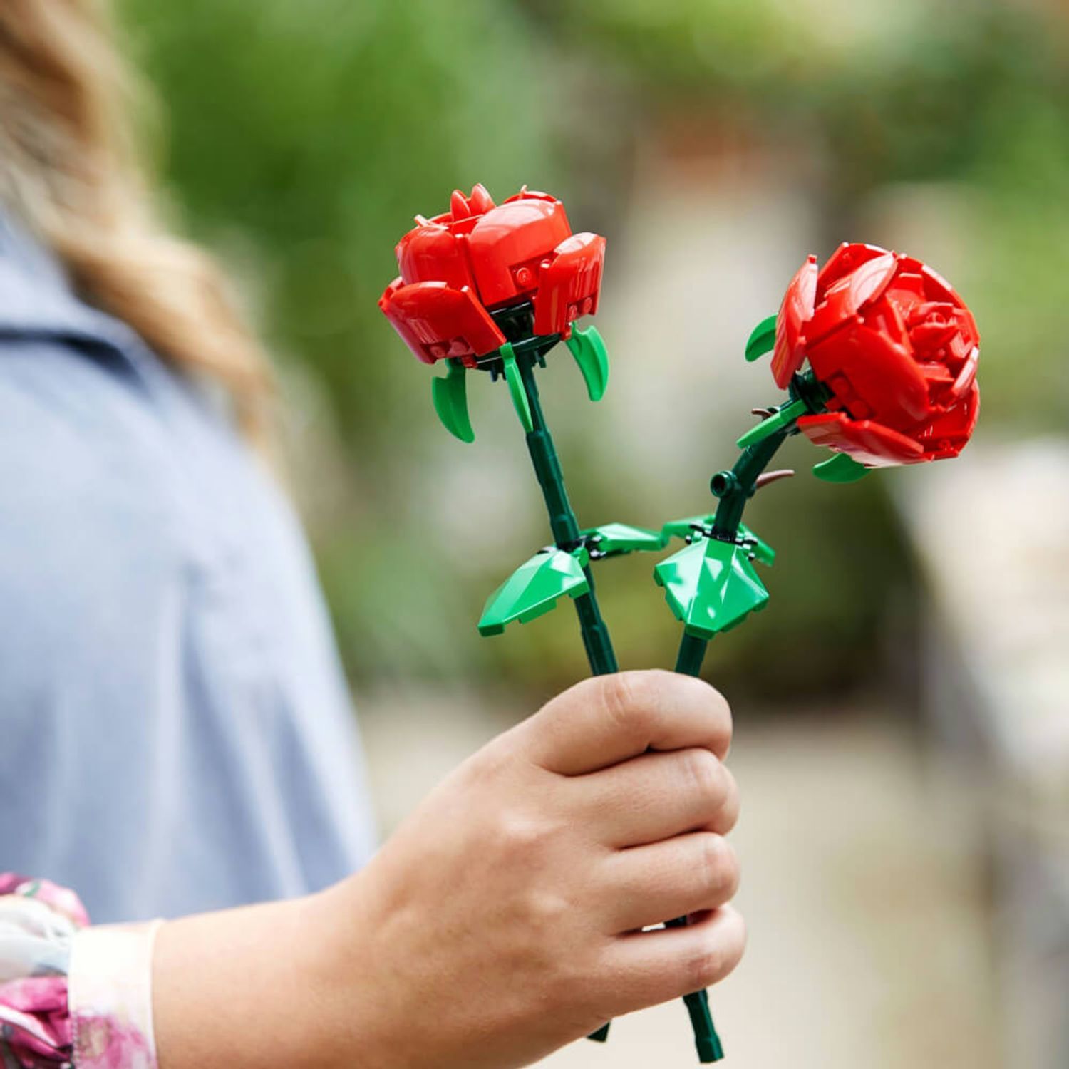 Due rose LEGO rosse in mano, foglie e steli verdi, su sfondo sfocato.