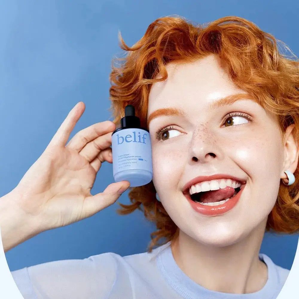 Donna con capelli rossi tiene in mano un flacone di siero Belif Super Drops su sfondo blu.
