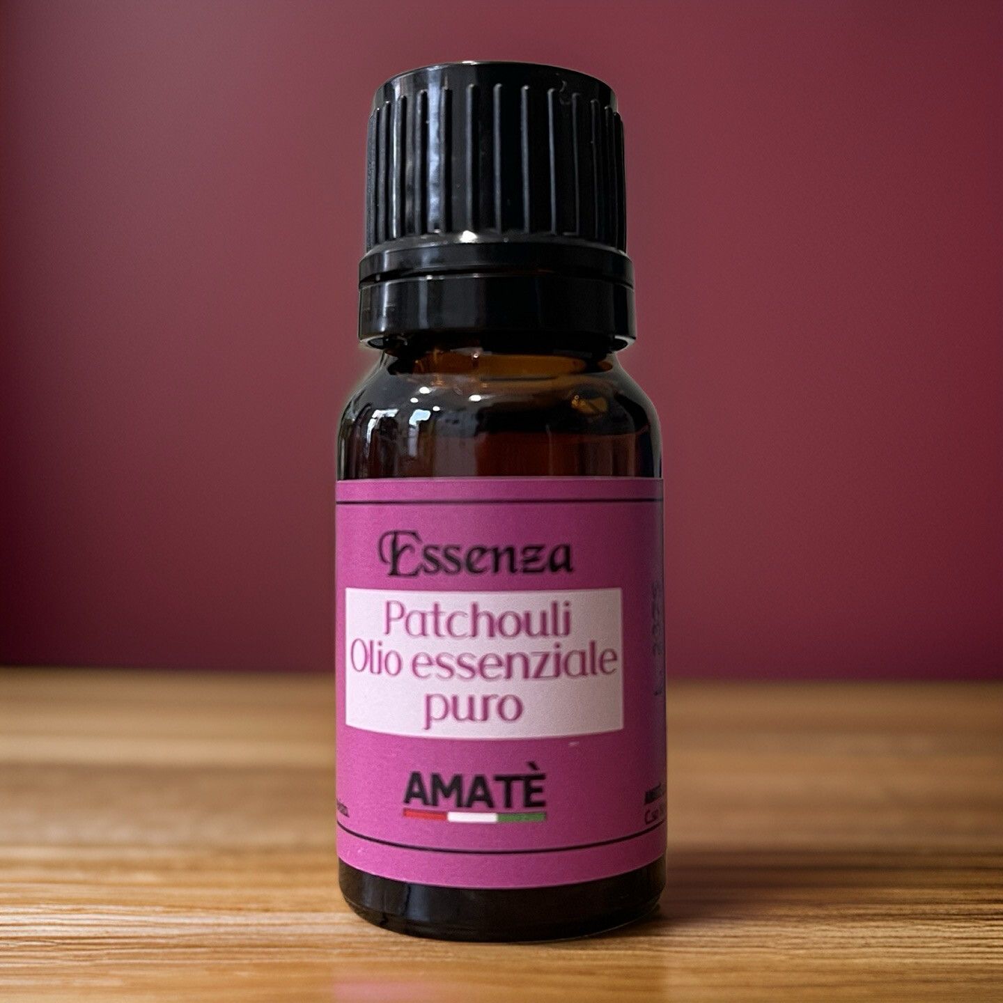 Flacone di vetro marrone con tappo nero. Etichetta con testo: Essenza Patchouli Olio essenziale puro. Marchio: AMATÈ. Su superficie in legno.
