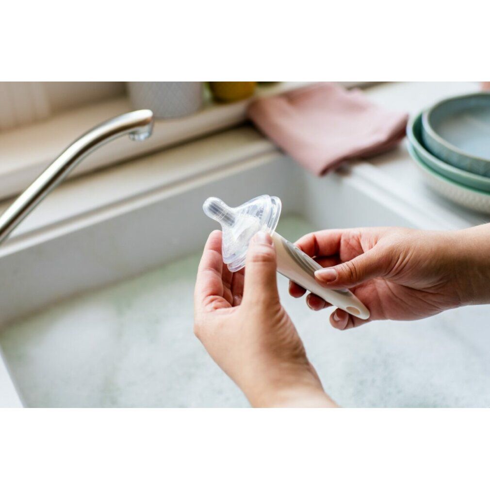 Mani che tengono una tettarella trasparente e un collo di bottiglia sopra un lavandino. Un rubinetto e stoviglie sullo sfondo.