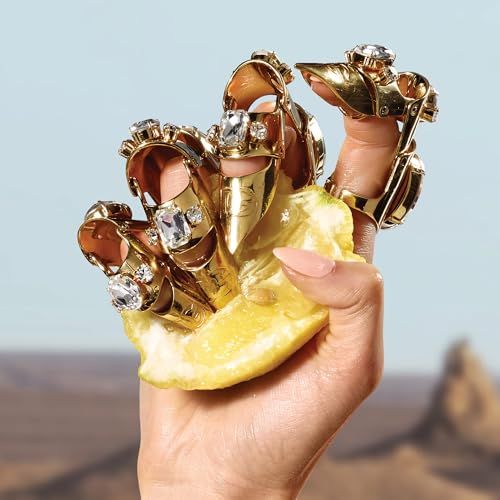 Mano con anelli dorati con pietre, tiene fetta di limone. Sfondo: deserto.