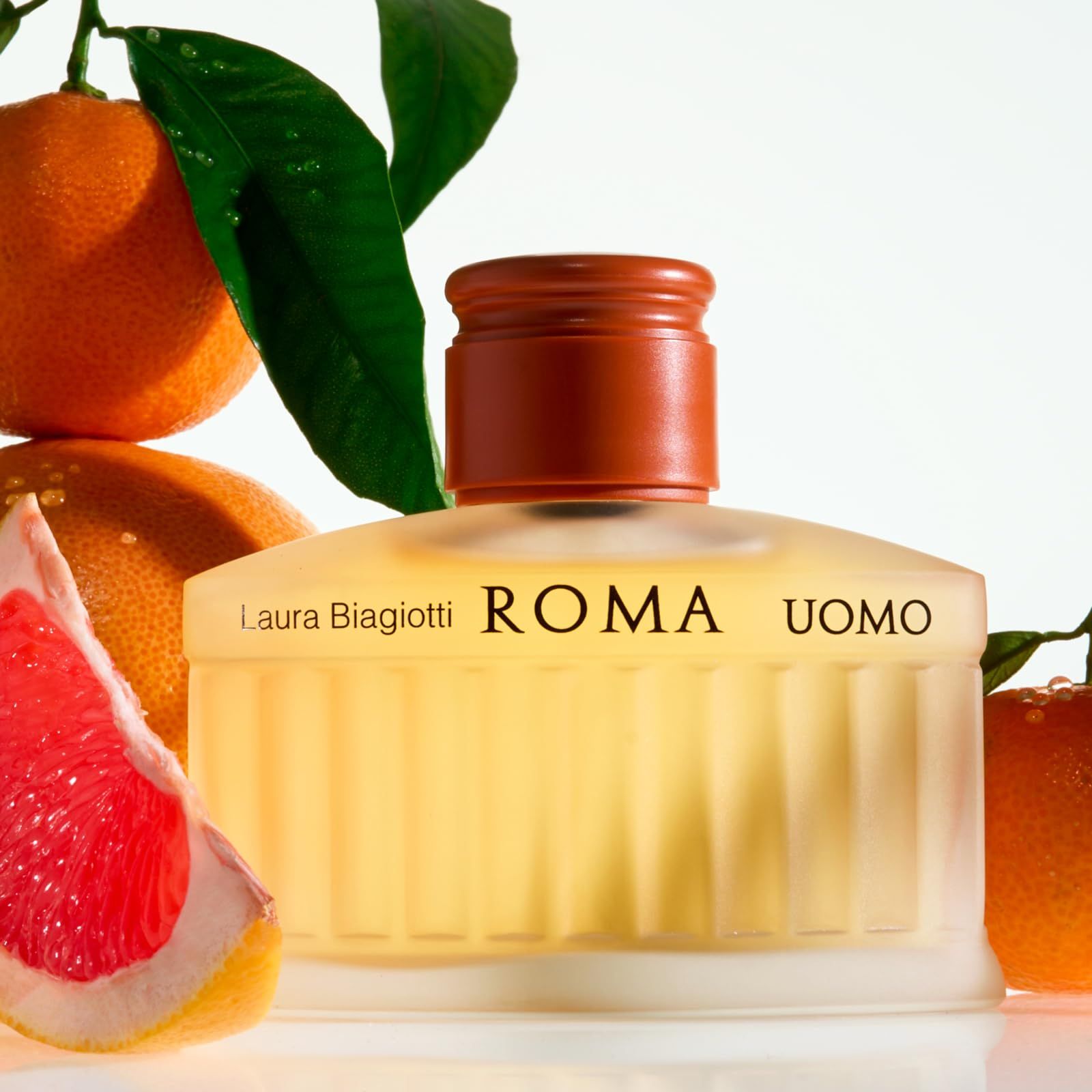 Flacone di profumo con liquido giallastro. Scritta: Laura Biagiotti ROMA UOMO. Circondato da arance e pompelmo.