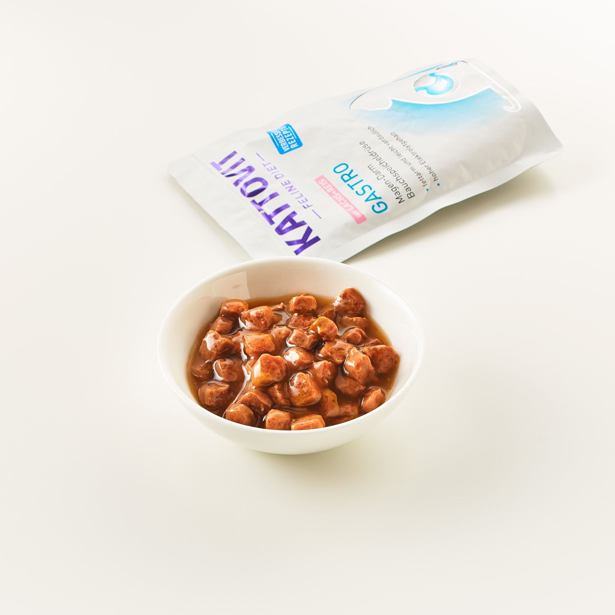 Ciotola con cibo accanto a busta aperta. Pezzi di cibo in salsa. Busta con logo KATTOVIT e nome prodotto GASTRO. Sfondo bianco.