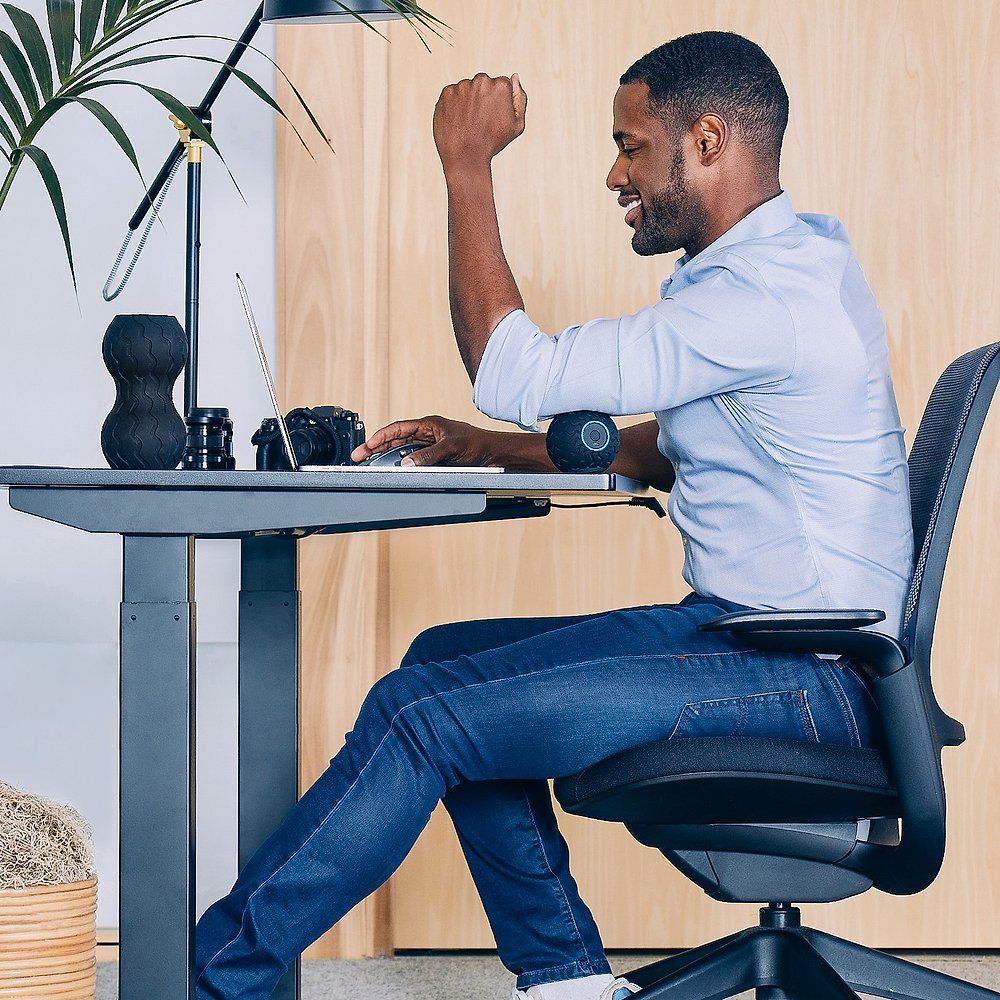 Uomo seduto a una scrivania, la sfera da massaggio sul braccio. Alza il pugno. Laptop e lampada visibili.