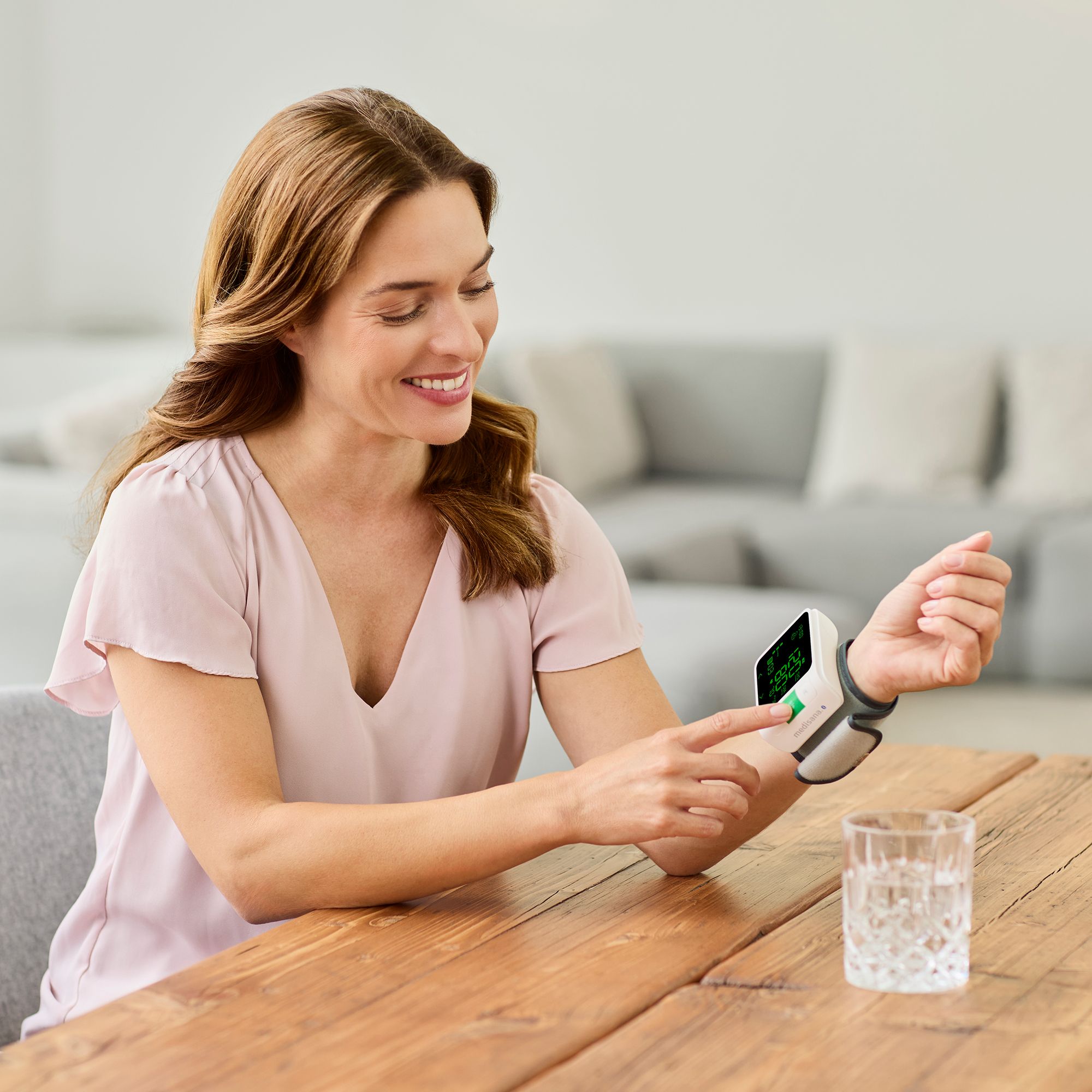 Una donna misura la pressione sanguigna al polso con un dispositivo medisana. Un bicchiere d'acqua è sul tavolo.