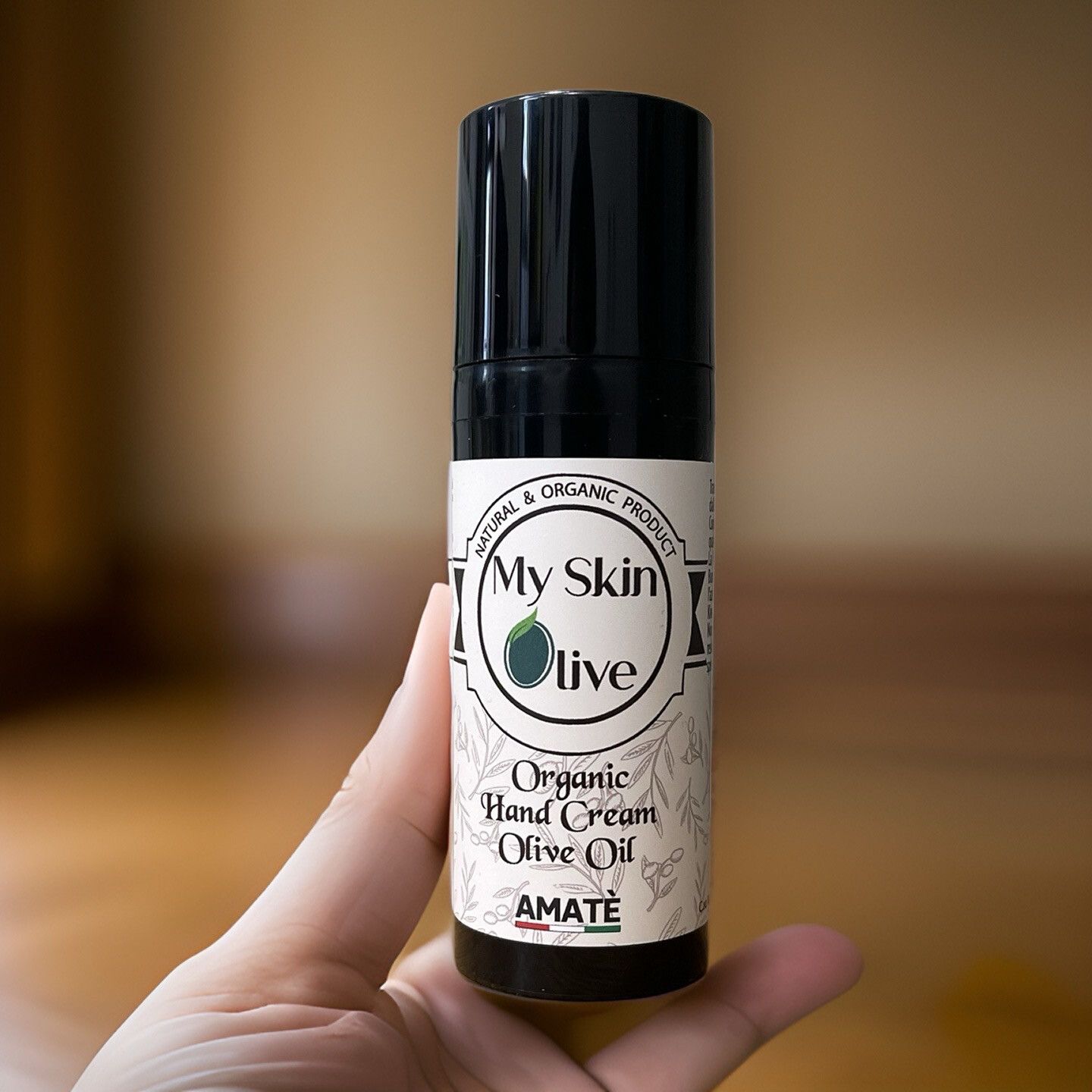 Flacone nero tenuto in mano. Scritta: My Skin Olive, Organic Hand Cream Olive Oil, Amatè.
