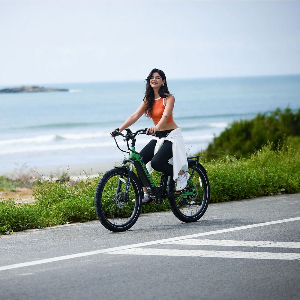 Donna in sella a un'e-bike verde su strada. Mare e costa sullo sfondo. Marchio Cyrusher visibile.