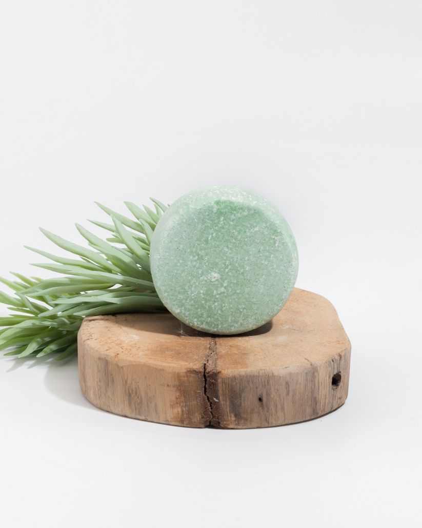 Panetto shampoo verde rotondo su un ceppo di legno. Fogliame verde accanto. Su sfondo bianco.