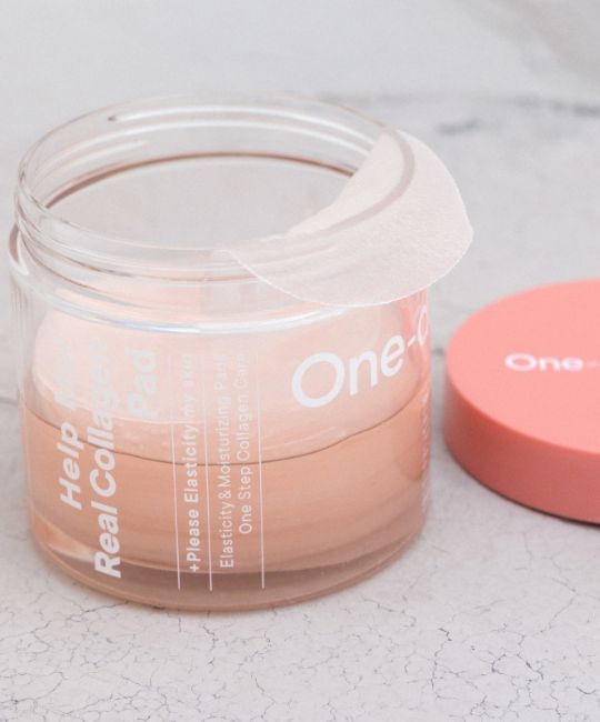 Barattolo trasparente aperto con coperchio rosa. Scritta: Help Me Real Collagen Pad. Tamponi visibili.