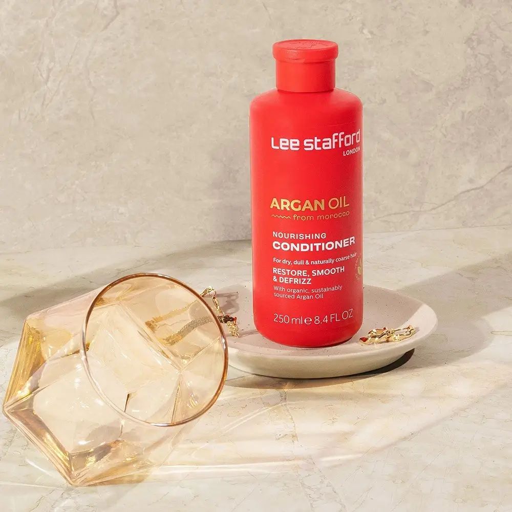 Flacone rosso di balsamo per capelli. Scritta: Argan Oil, Nourishing Conditioner. Su un piatto, vicino a un bicchiere.