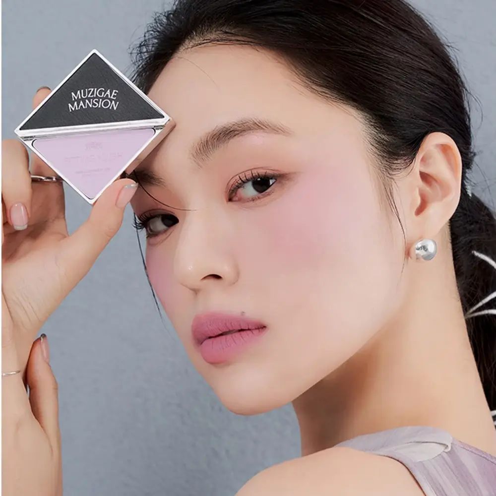 Fard in forma di diamante, tenuto da una persona. Marchio MUZIGAE MANSION visibile. Tonalità rosa. Trucco sul viso.