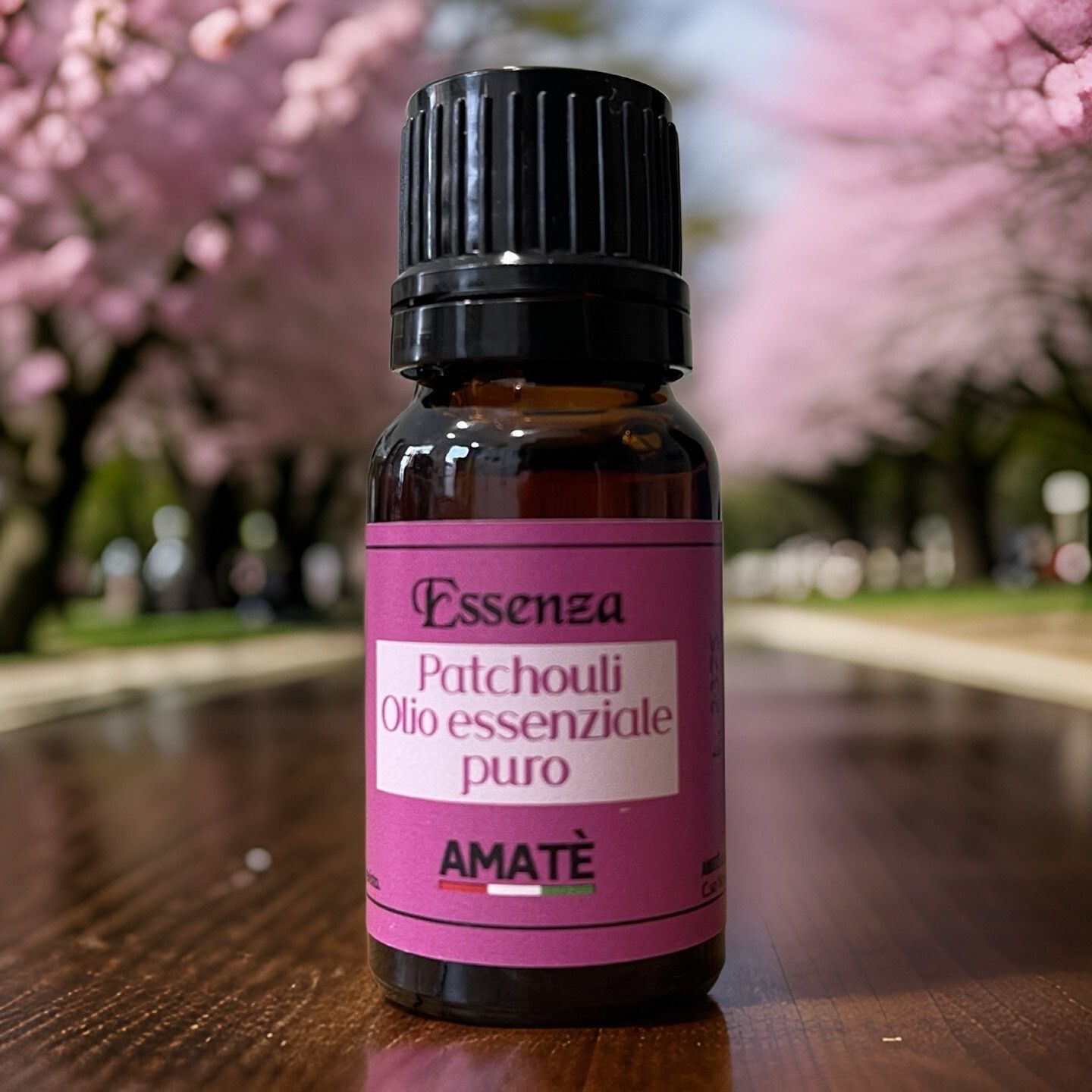 Flacone di vetro marrone con tappo nero. Etichetta con testo: Essenza Patchouli Olio essenziale puro. Marchio: AMATÈ. Sfondo: alberi.