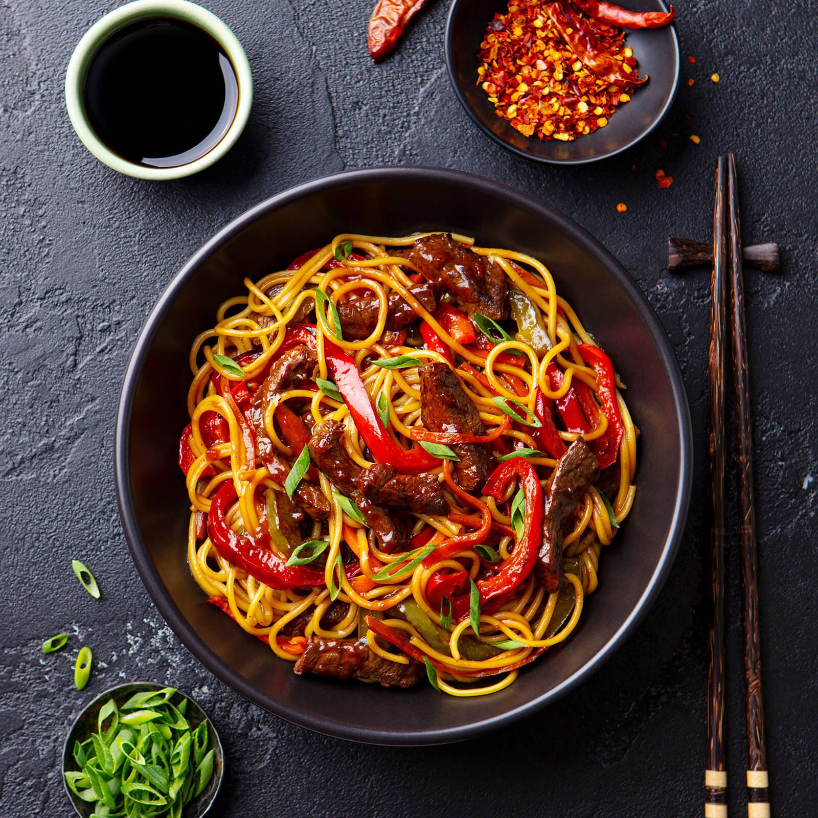 Piatto nero di spaghetti con carne, peperoni e salsa. Bacchette accanto.