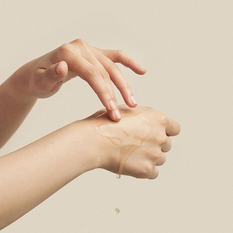 Mani che applicano un liquido sulla pelle. Liquido che gocciola. Pelle chiara, sfondo beige.