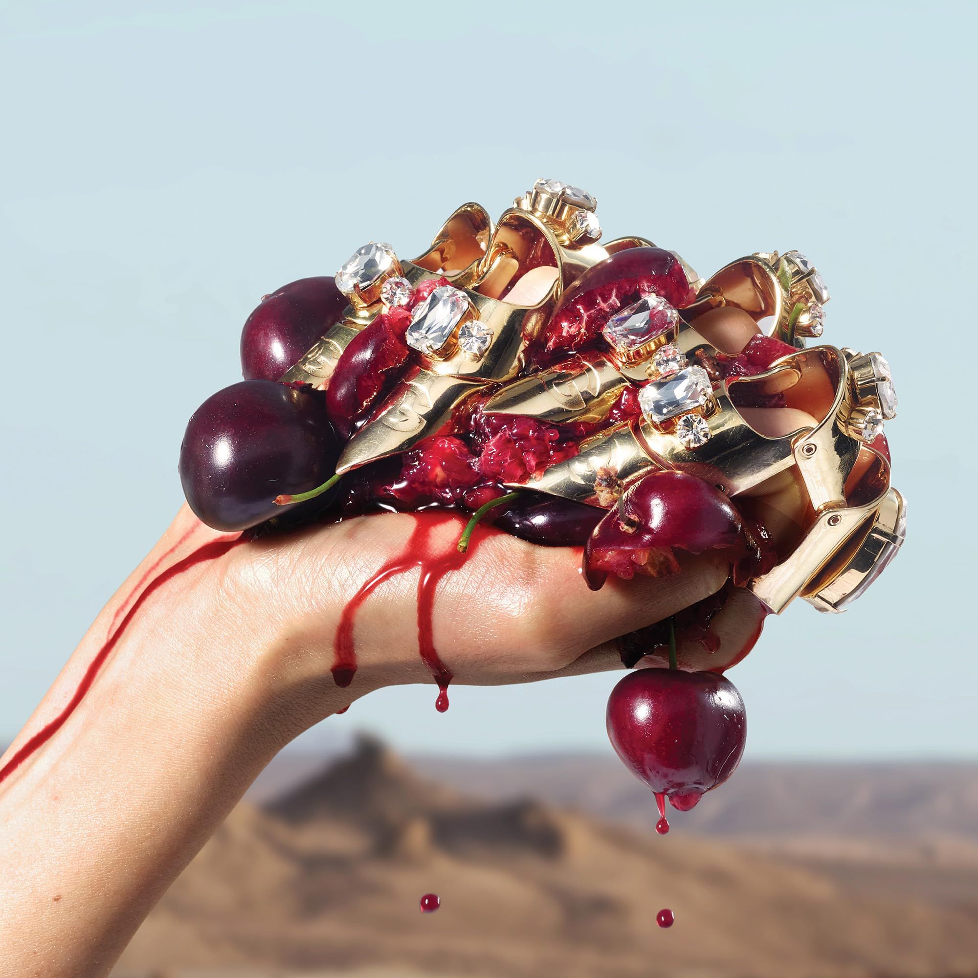 Mano con anelli dorati con pietre preziose. La mano tiene ciliegie e succo di ciliegia. Sfondo: deserto e cielo blu.