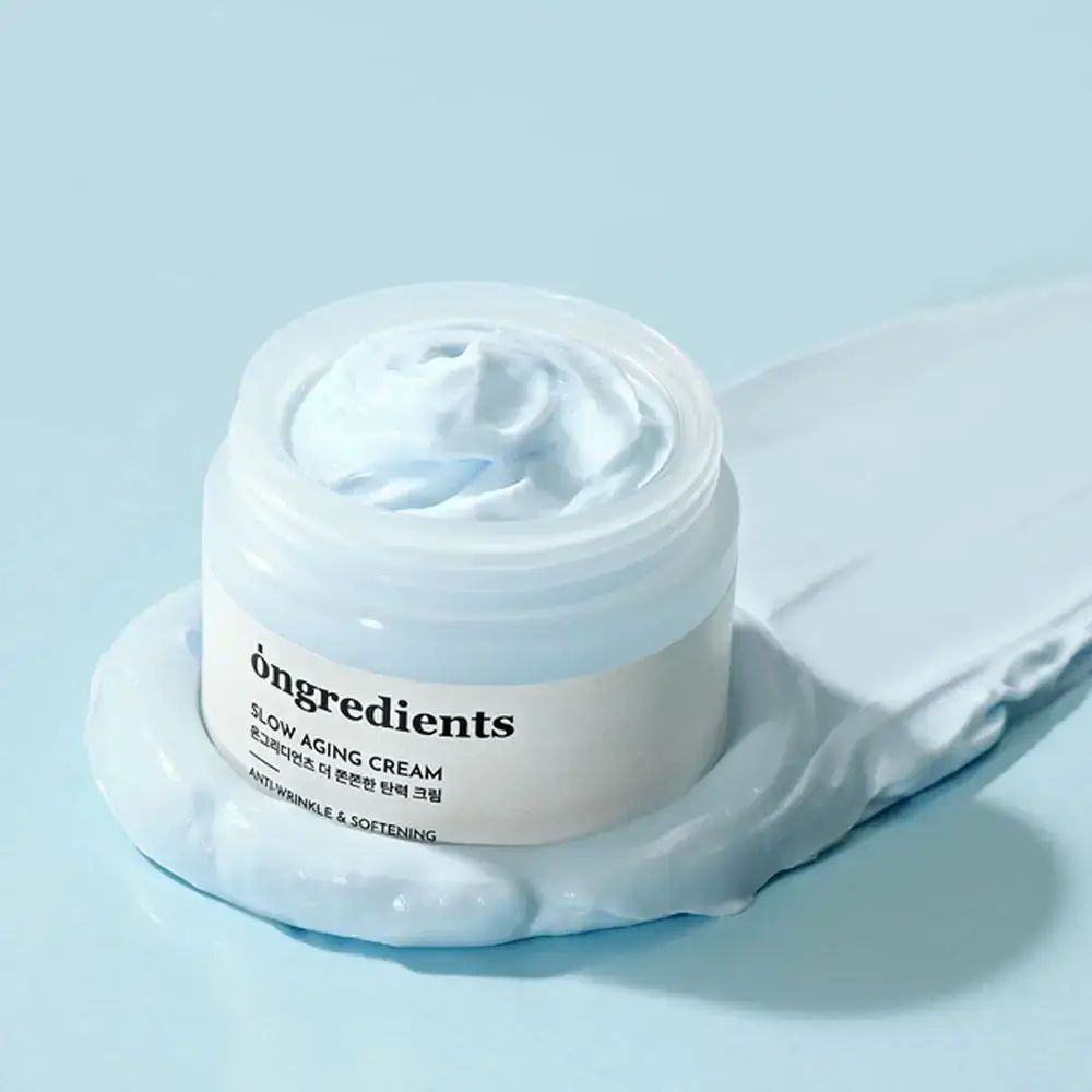Crema in vasetto trasparente. Scritta: ONGREDIENTS Slow Aging Cream. Texture su sfondo azzurro.