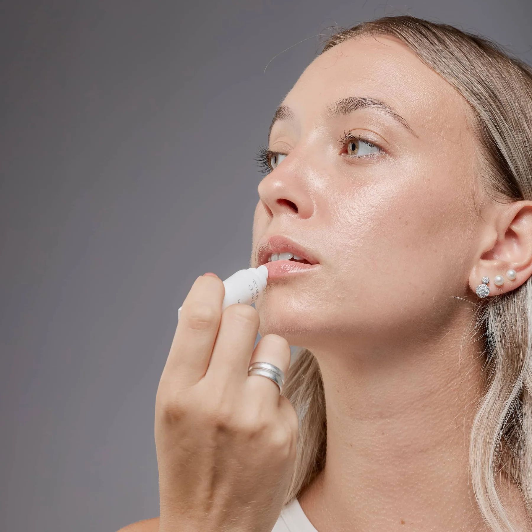 Una donna applica il balsamo per le labbra. Il tubo è bianco. Indossa un anello e orecchini. I suoi capelli sono biondi.