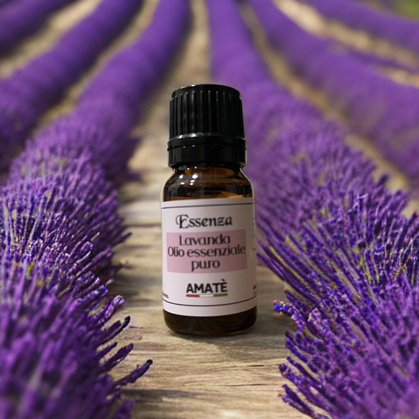 Flacone di vetro marrone con tappo nero, davanti a campi di lavanda. Etichetta con testo: Essenza Lavanda Olio essenziale puro. Marchio: AMATÈ.