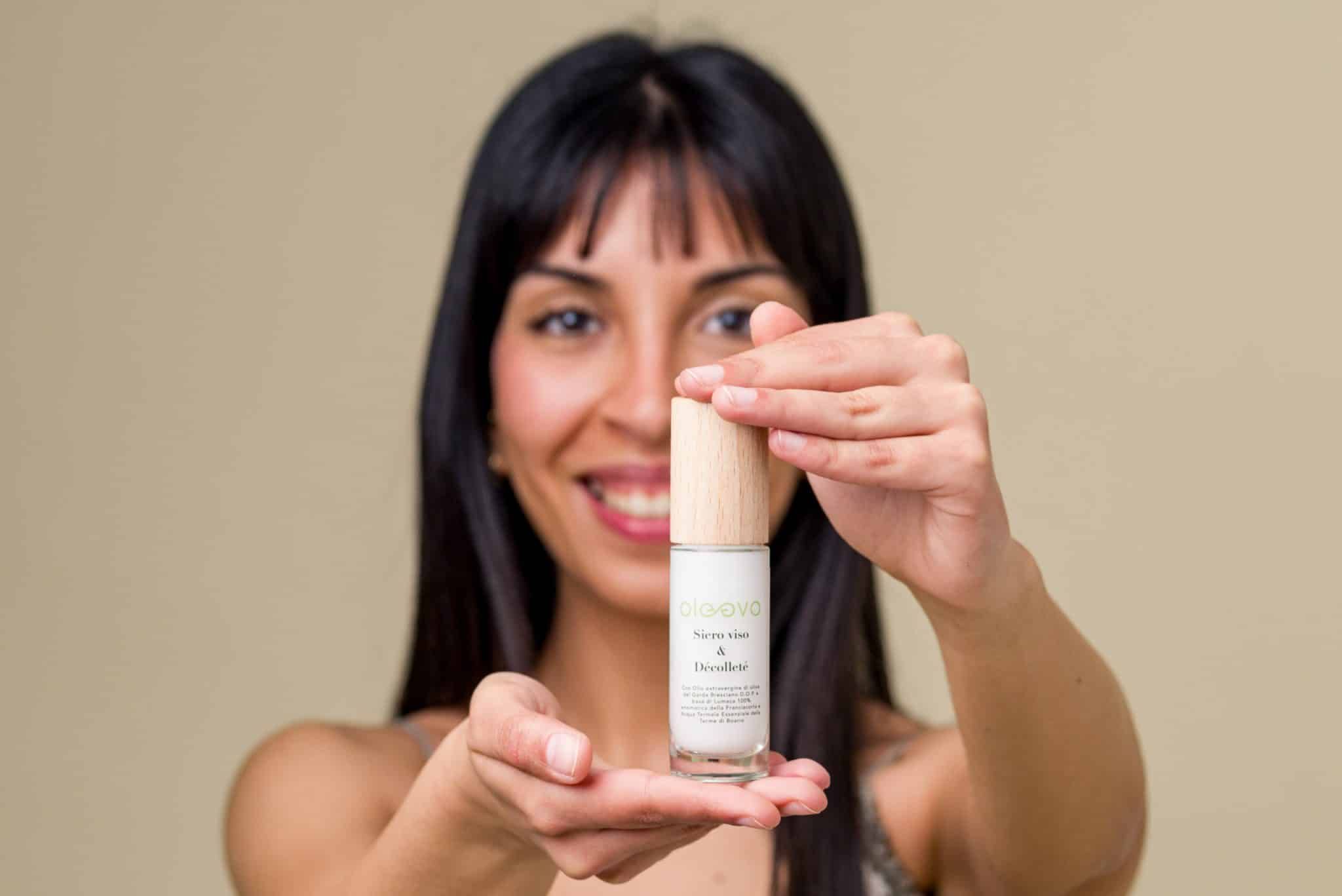 Donna tiene in mano un flacone di siero. Scritta: Oleevo, Siero viso & Décolleté. Sfondo chiaro.