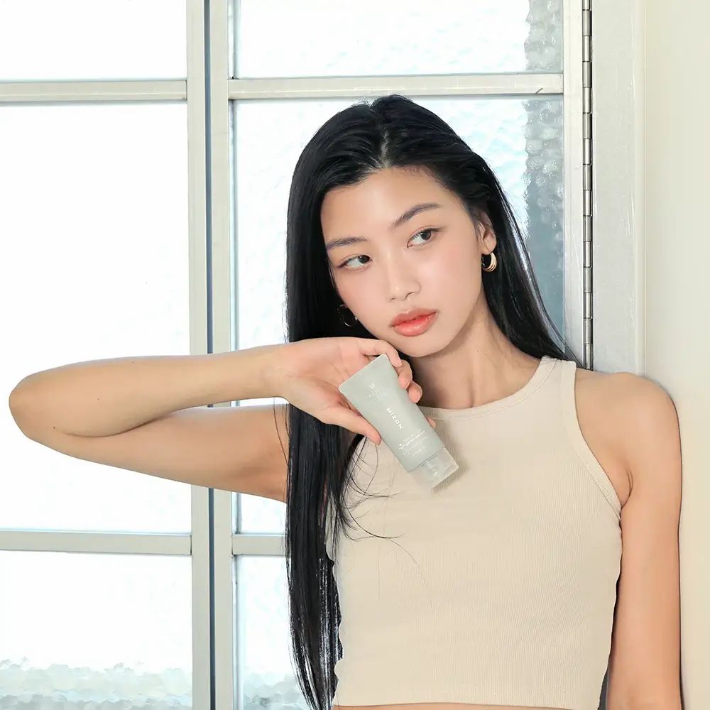 Donna tiene un tubetto grigio. Nome del prodotto non visibile. Indossa una canotta.