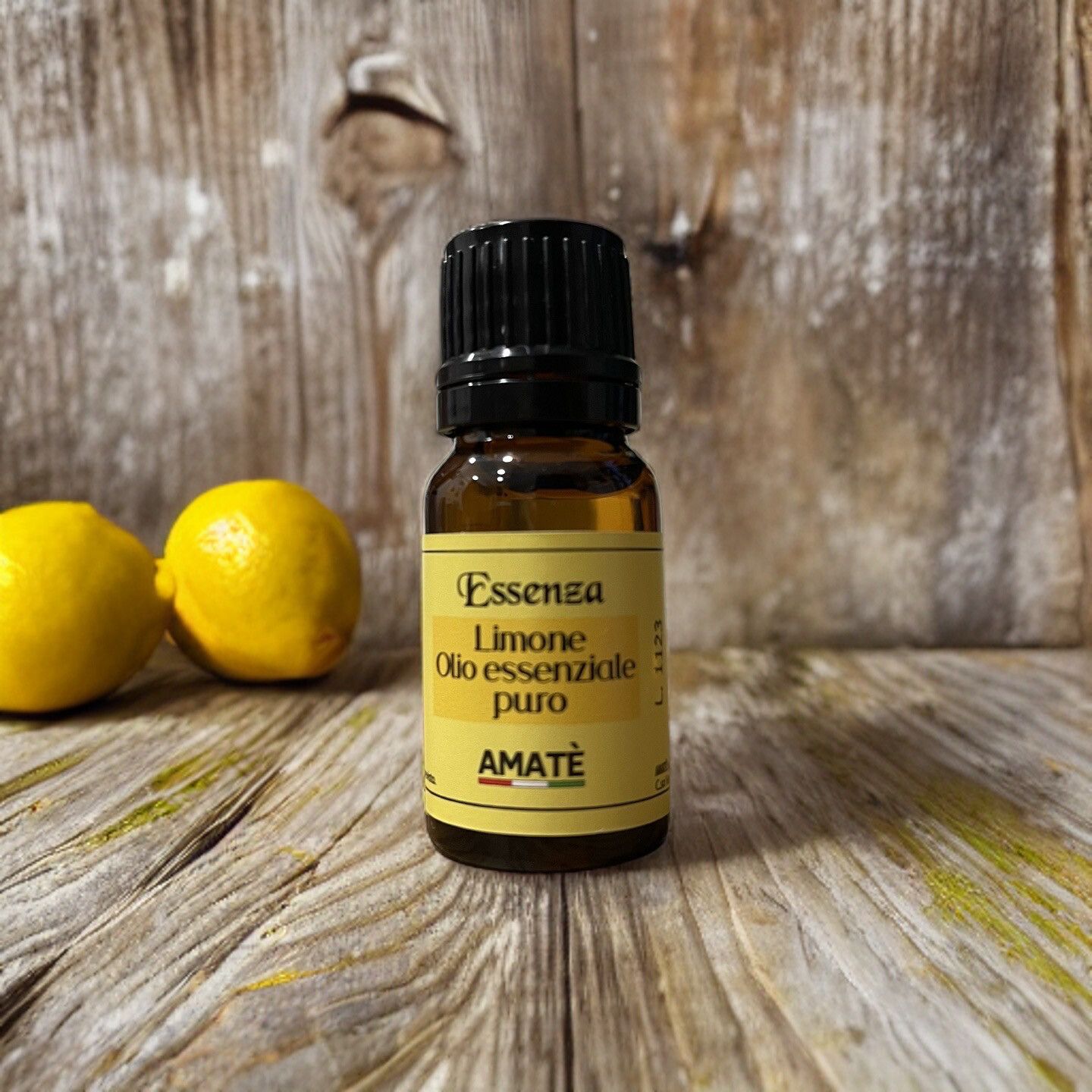 Flacone di vetro marrone con tappo nero, davanti a sfondo in legno con limoni. Etichetta gialla con testo: Essenza Limone, Olio essenziale puro. Logo AMATÈ.