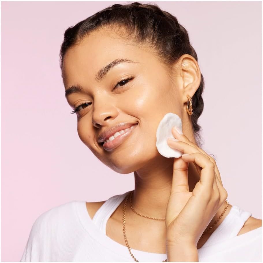 Una donna pulisce il viso con un dischetto di cotone. Indossa una maglietta bianca e ha i capelli intrecciati.