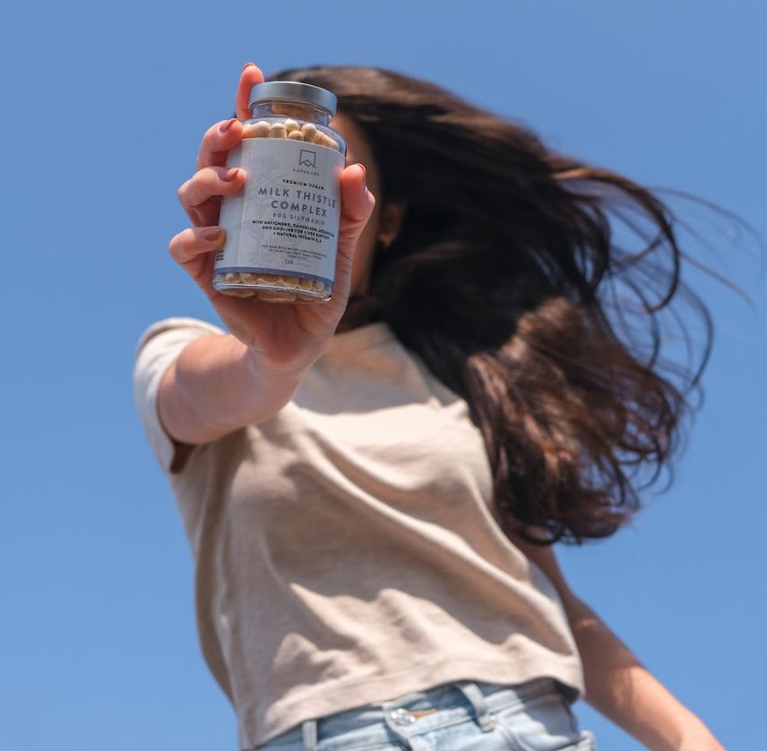 Persona che tiene in mano un flacone di capsule. Scritta: Milk Thistle Complex. Cielo blu.