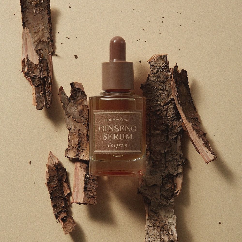 Flacone di siero con tappo marrone ed etichetta. Circondato da pezzi di legno. L'etichetta riporta "Ginseng Serum" e "I'm from".