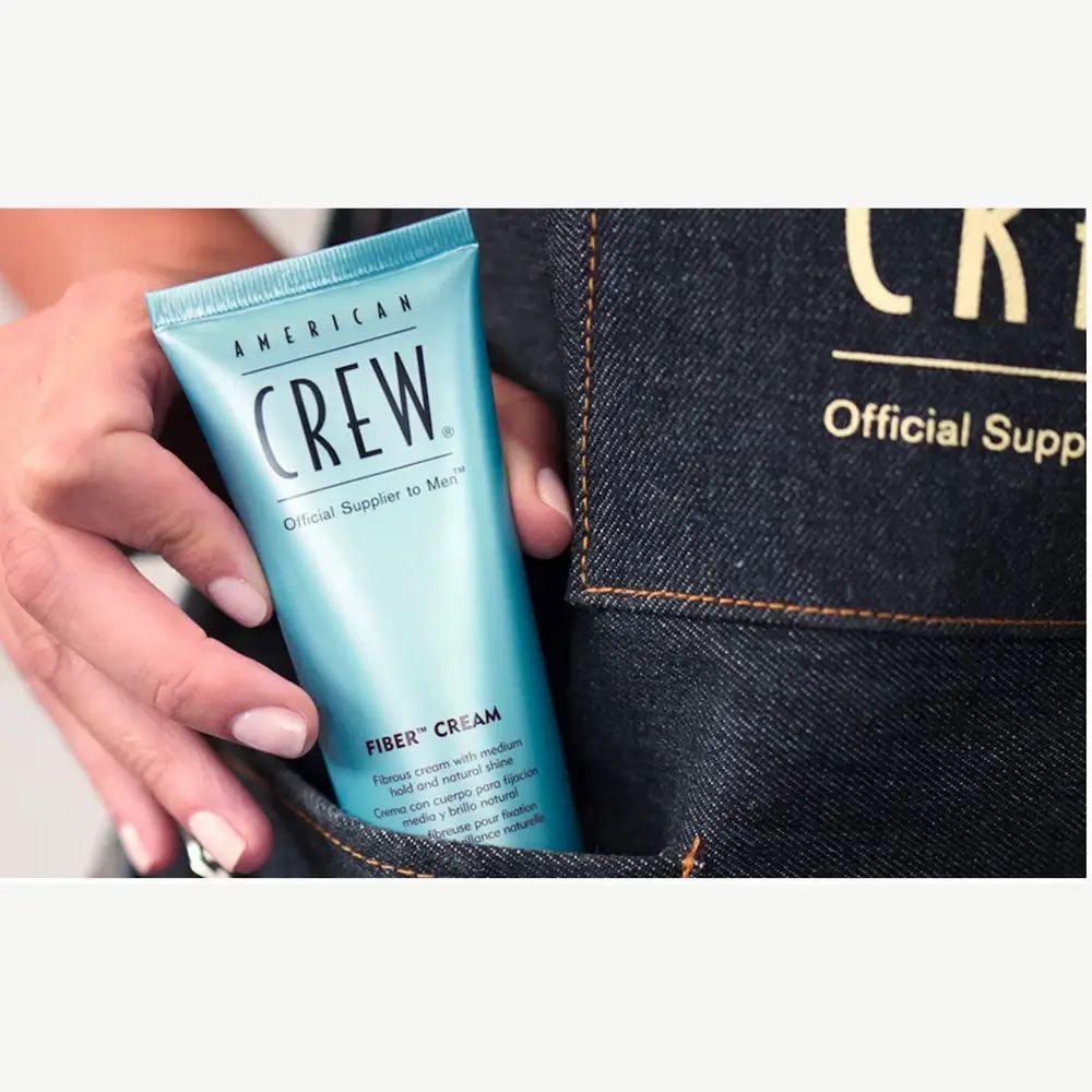 Tubo blu chiaro in una mano. Scritta: American Crew, Fiber Cream. Su sfondo jeans.
