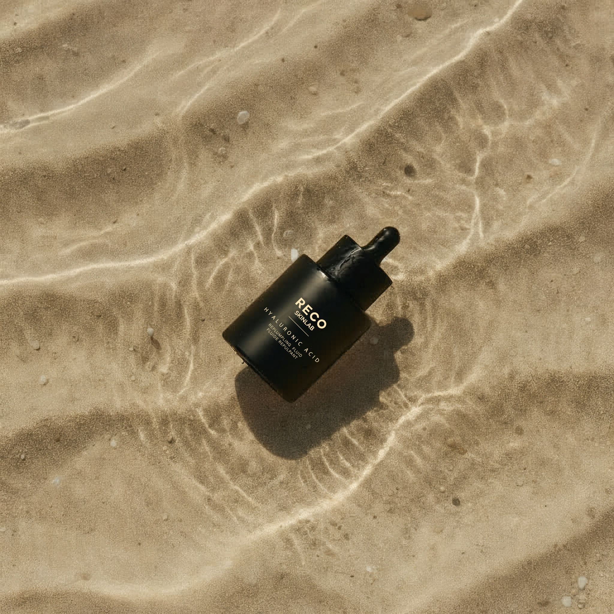 Flacone nero con pipetta nell'acqua. Scritta: RECO SKINLAB, Acido Ialuronico. Acqua con fondo sabbioso.