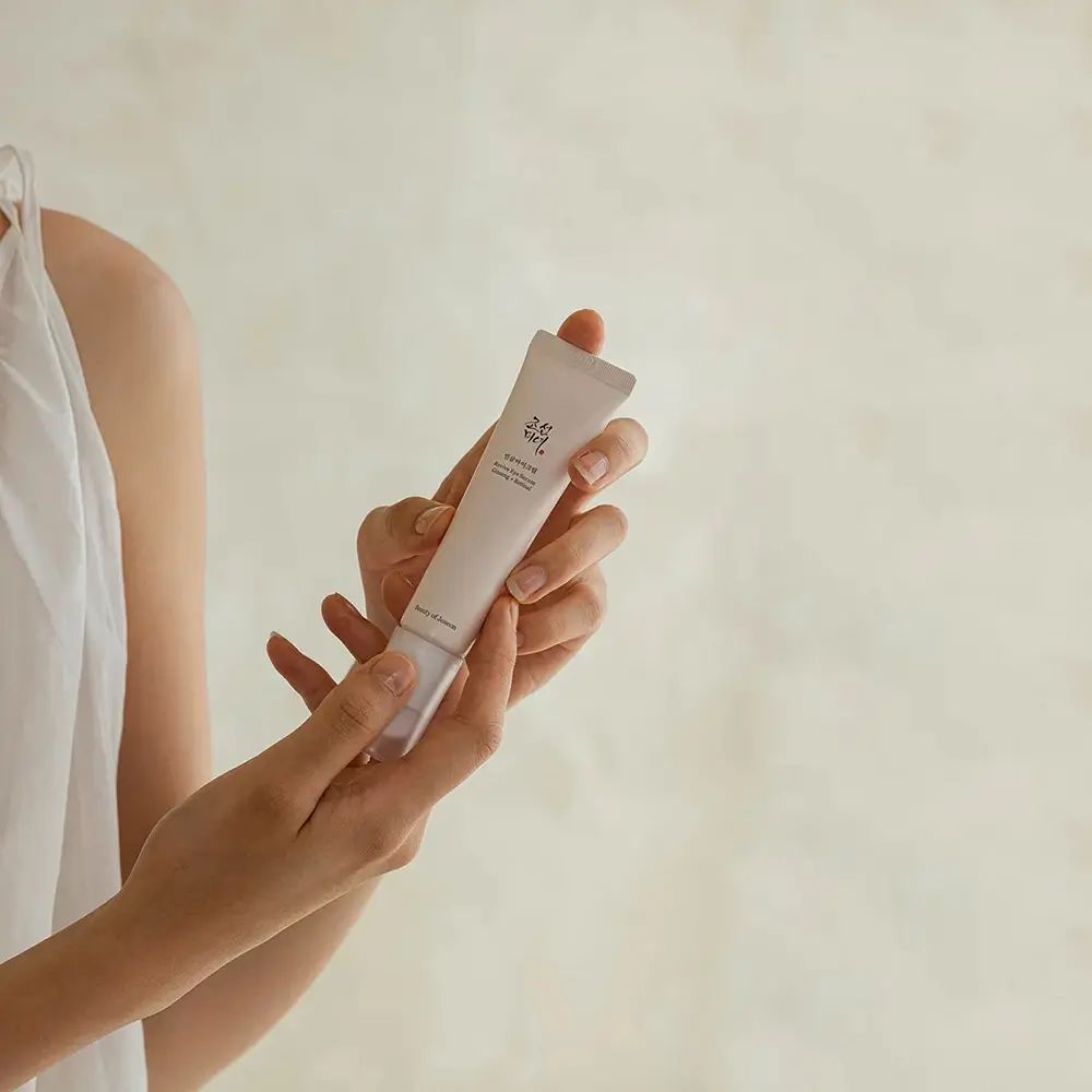 Mani che tengono un tubo di crema. Tubo rosa chiaro con tappo bianco. Marchio: Beauty of Joseon.