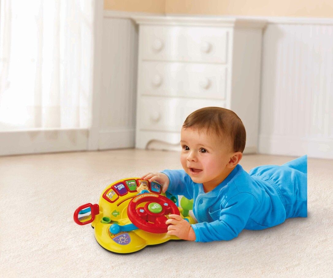 Bambino sdraiato a pancia in giù che gioca con un volante giocattolo giallo. Il bambino sorride.