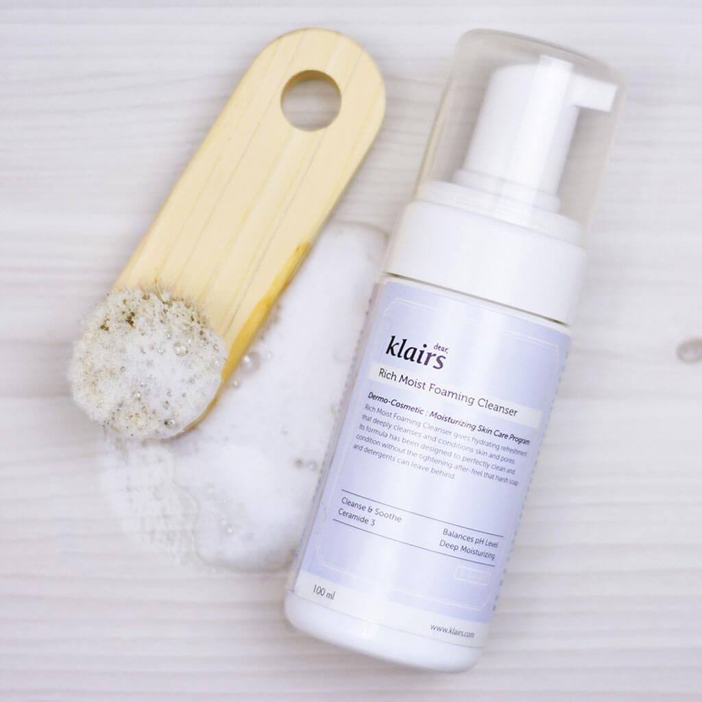 Flacone di schiuma bianco con logo Klairs e testo. Spazzola per il viso in legno accanto. Schiuma.
