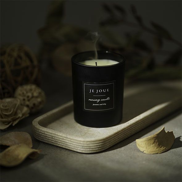 Candela nera fumante su un vassoio. Scritta: Je Joue, massage candle, Jasmine Lily. Decorazioni sullo sfondo.