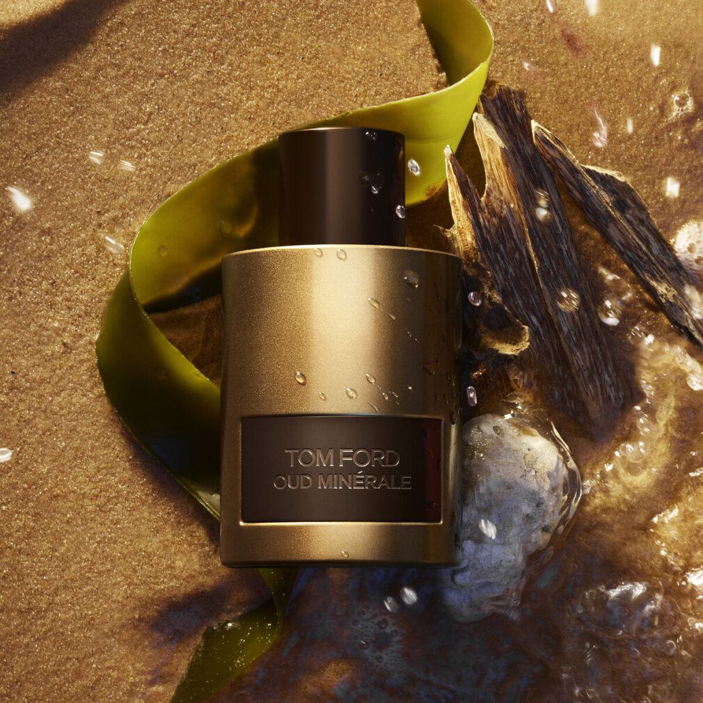 Flacone dorato, tappo marrone, etichetta rettangolare con testo TOM FORD OUD MINERALE. Su sabbia, con acqua.