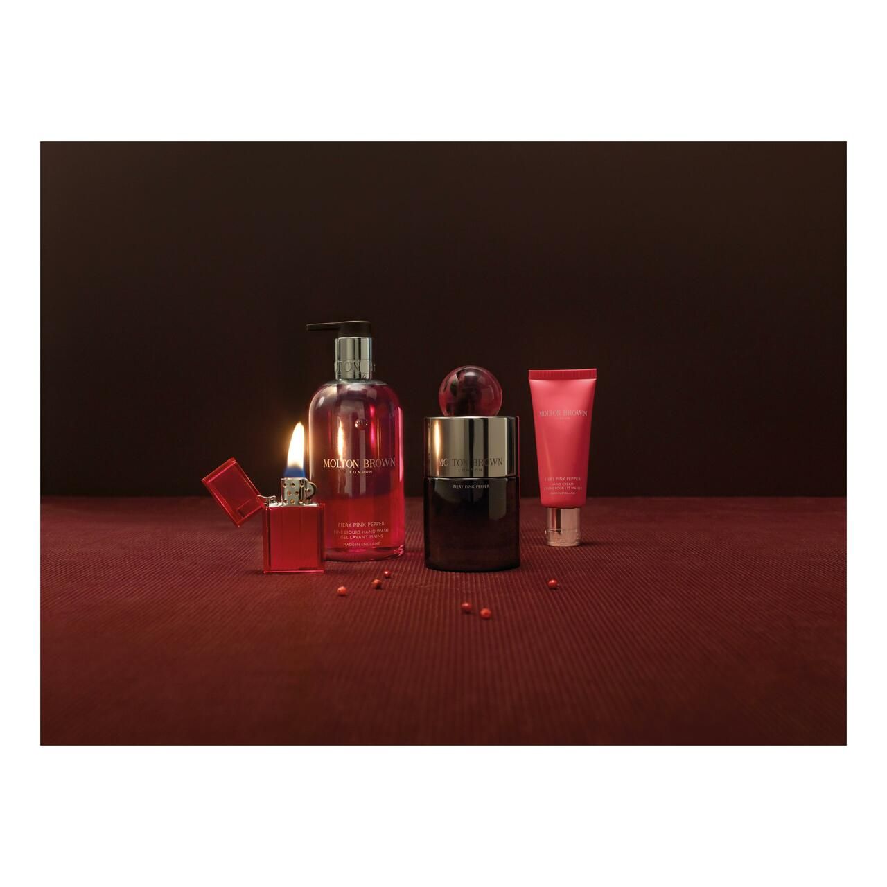 Disposizione di prodotti su sfondo rosso. Include flacone, flacone, tubetto di crema e accendino. Marchio: Molton Brown.
