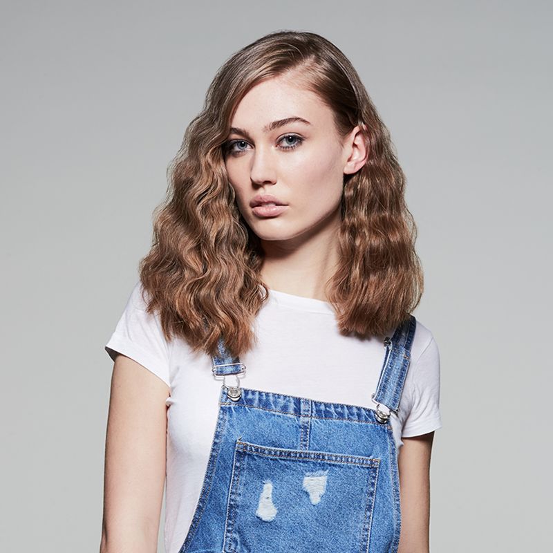 Donna con capelli mossi. Indossa una maglietta bianca e una salopette di jeans. Sfondo grigio.