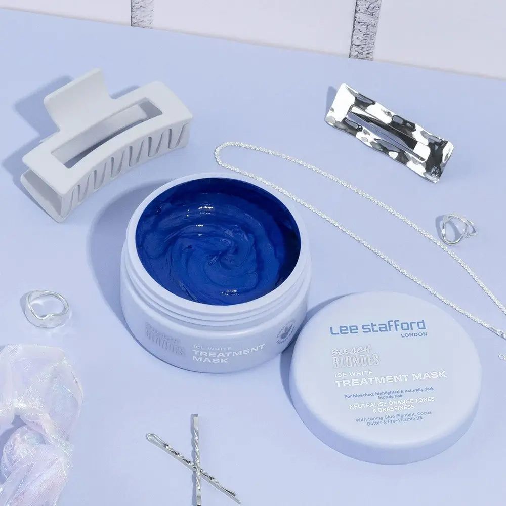 Barattolo bianco aperto con maschera blu, accanto a fermagli per capelli e gioielli su sfondo chiaro.